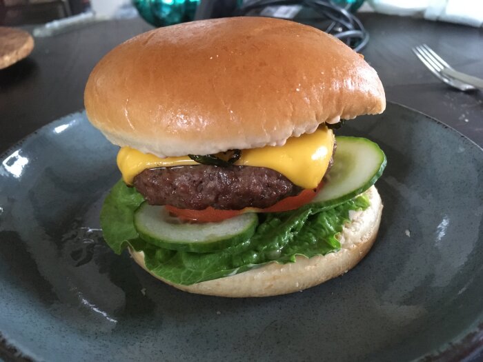 En cheeseburger med sallad, tomat, gurka, ost och brödbulle på en mörk tallrik.