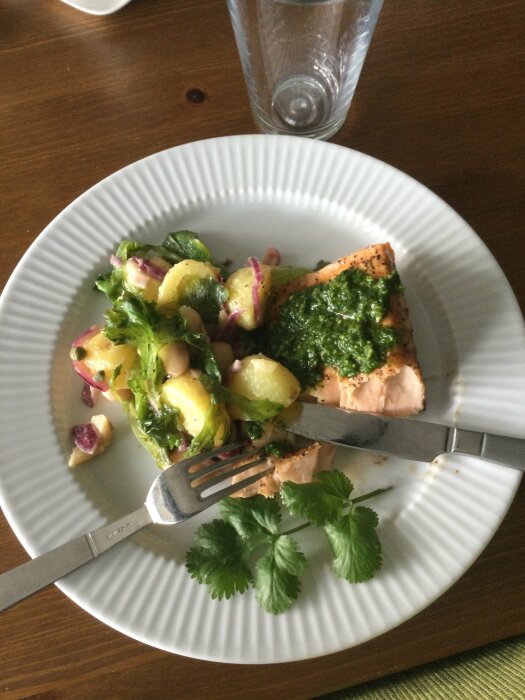 Tallrik med lax, potatissallad, pesto, bestick på träbord, glas vatten, fräscha örter, måltid, hälsosam, ljusinfall.