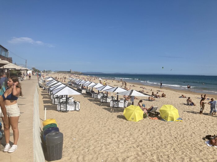 Solig stranddag, människor solar och badar, parasoll radade, promenadväg bredvid, klart himmel.