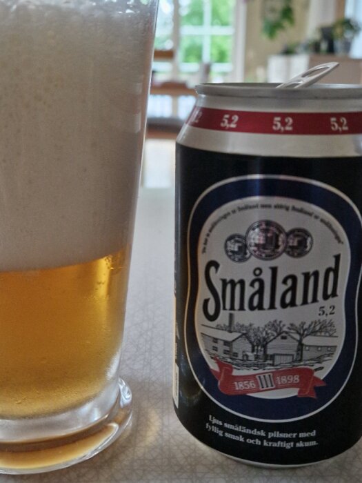 Ölglas med skum, burk av Småland öl framför fönster, 5.2% alkohol, svensk pilsner.