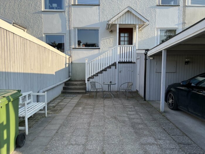 Tvåvåningshus, trappa, balkong, garage, bil, uteplats, soptunna, vit bänk, fönster, eftermiddagssol.