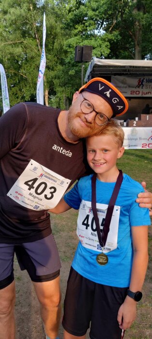 Två leende löpare, en vuxen och ett barn, med nummerlappar och medalj, firar efter ett lopp.