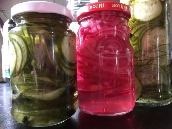Tre glasburkar med inlagda grönsaker, en med röd lösning, på en träyta.
