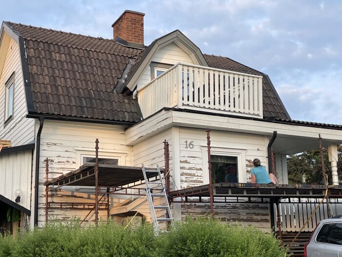 Renoveringsarbete på ett vitt hus med tegeltak, byggställningar, personer på veranda, nummer 16, grönska.