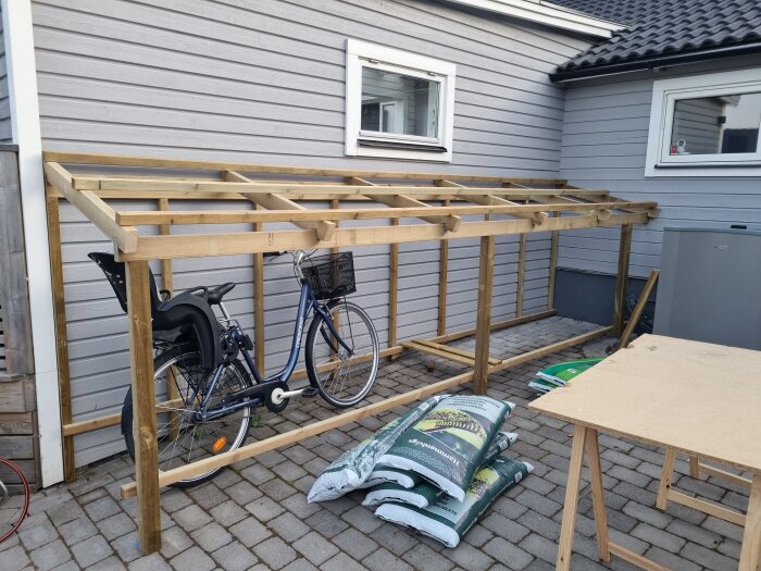 Träkonstruktion för tak, ofärdigt byggprojekt, cykel, säckar, husvägg, fönster, stenläggning.