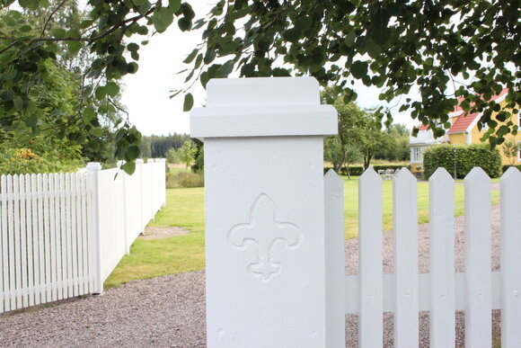 Vitt staket och stolpe med dekorativt emblem, grusväg, grönt gräs och träd, lantligt landskap i bakgrunden.