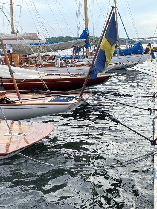 Svenska flaggor på segelbåtar vid brygga, reflektion av vatten, molnig himmel, båtens skrov synligt.