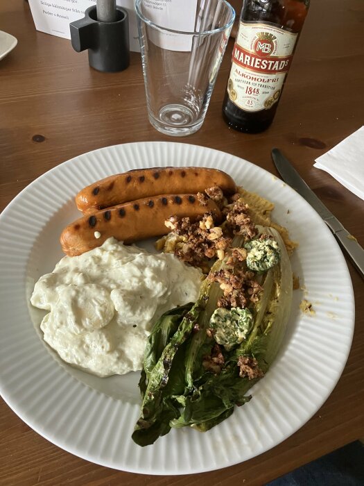 Måltid med korv, potatissallad, grillad sallad, tomater, ölflaska "Mariestads alkoholfri", tomt glas, mörkt bord.