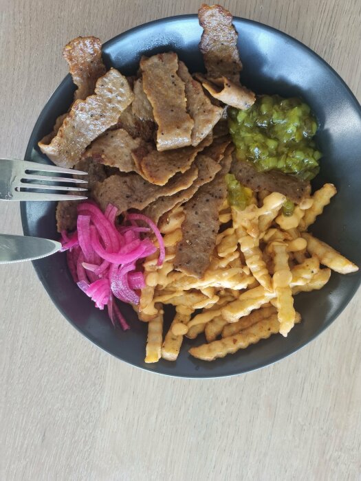 En tallrik med kebabkött, pommes frites, löksallad och guacamole på ett träbord.