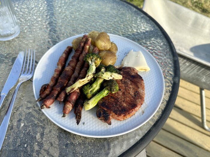 Grillad hamburgare, baconlindade sparris, rostad broccoli, potatis, på en utomhusglasbord, soligt, måltidspresentation.