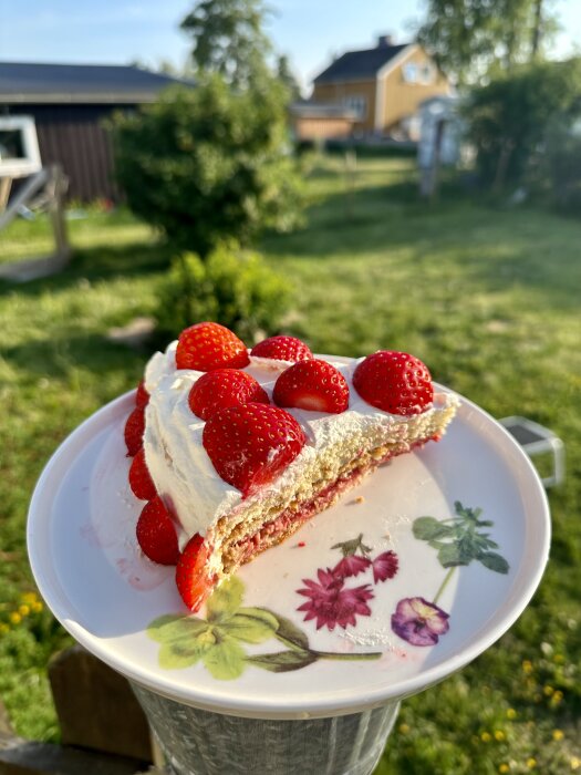 Ett stycke jordgubbstårta på tallrik i trädgård, suddig bakgrund med hus och grönska, sommardag.
