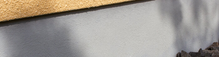 Del av beige yttre vägg, textur av puts, skuggor, stenar nederst, naturlig belysning, arkitektonisk detalj.