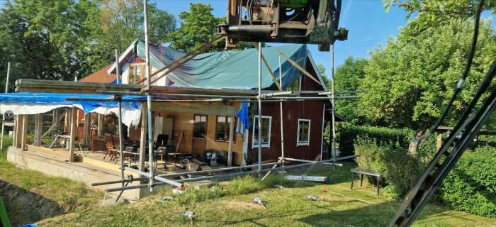 Hus under renovering, ställningar, presenningar, grönska, träd, ingen synlig personal, dagsljus, byggprojekt på landsbygd.