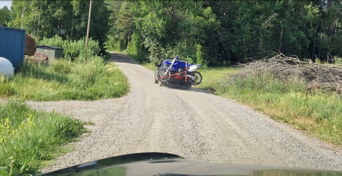 Motorcykel på väg, fastsatt på en släpvagn, på en grusväg i lantlig miljö. Solljus, grönska, sommar.