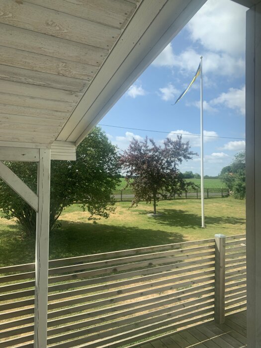 Altan med utsikt över en trädgård, en svensk flagga i vinden, gröna träd och blå himmel med moln.