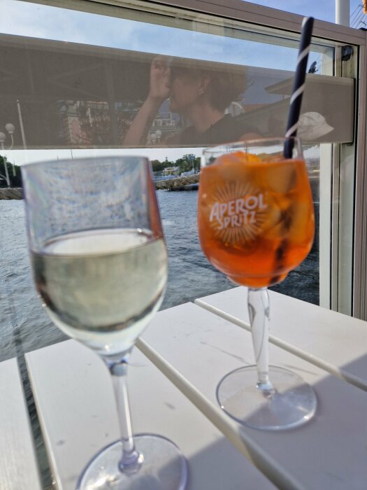 Glass vin, Aperol Spritz, reflekterande person i fönster, vattenutsikt, sugrör, avkopplande fikastund utomhus.