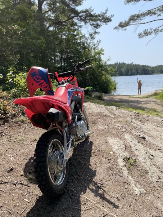 Motorcykel i fokus med naturen i bakgrunden; person i vattnet vid en sjö och soligt väder.