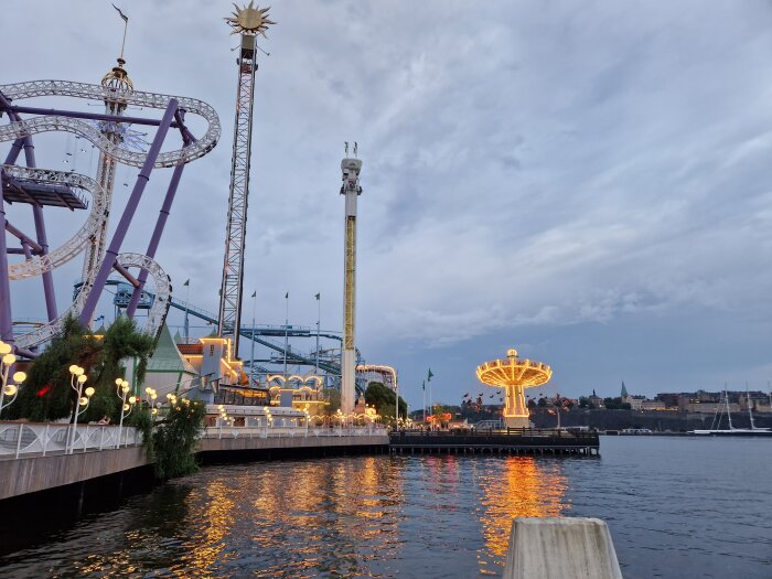 Nöjespark vid vattnet, berg-och-dalbana, attraktioner, kvällshimmel, upplyst karusell, reflektioner i vattnet, molnigt.