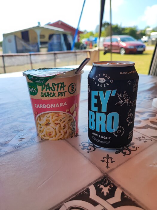 Mat på campingbord: Pasta Snack Pot, ölburk "EY BRO", utomhus, tält i bakgrunden, sommar.