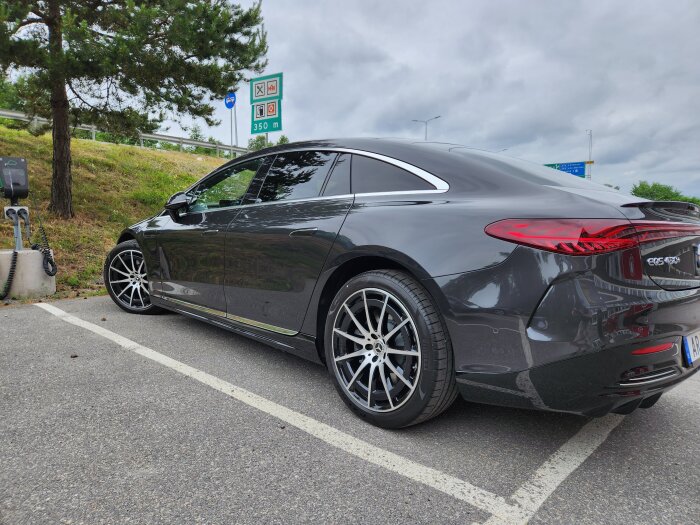 Svart sportig sedanbil parkerad vid laddstation under molnig himmel nära vägskyltar.