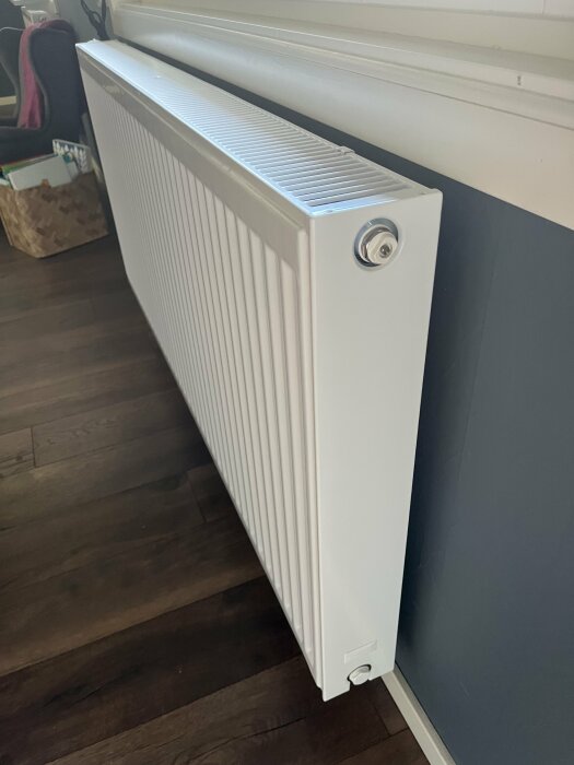 Vit element vid vägg, mörkt laminatgolv, inomhusmiljö, värme, radiator, justeringsknopp, grå vägg, energi.