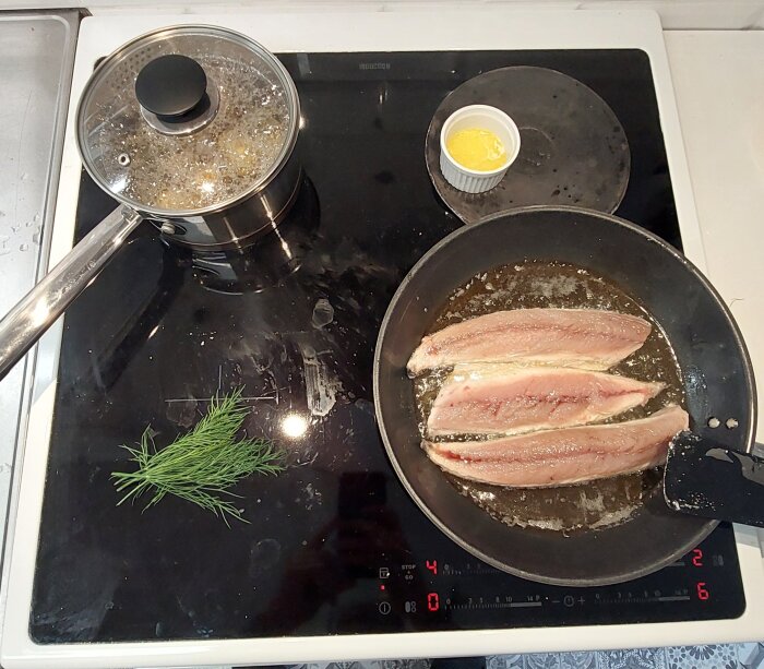 Två fiskfileer steks i panna; kastrull med lock, dillkvist och smält smör på induktionshäll.