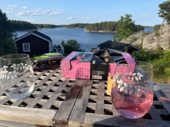 Uteservering med tomma glas, chokladkartong, sjöutsikt, liten stuga, trädäck, soligt, fridfullt.