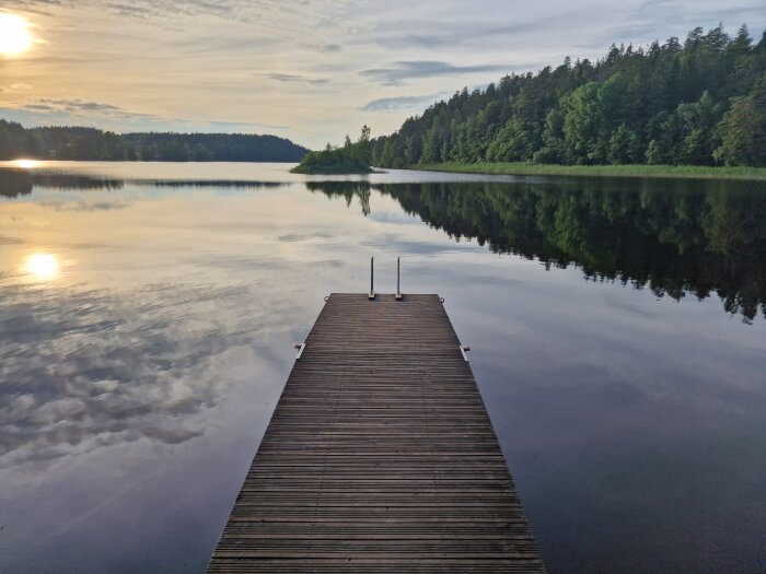 En träbrygga sträcker sig ut i en lugn, spegelblank sjö omgiven av skog och himlen återspeglas i vattnet.