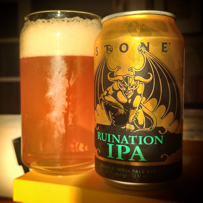 Ett glas med skummande öl bredvid en ölburk med etikett, "Ruination IPA". Warm belysning.