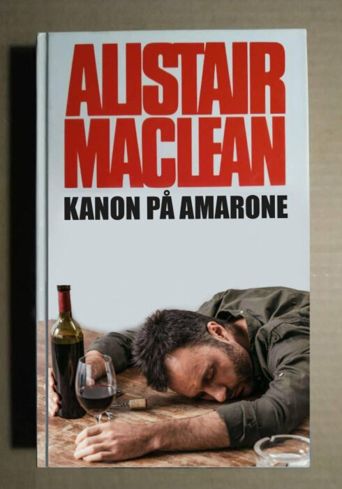 En bok med titeln "Alistair MacLean: Kanon på Amarone", en vinflaska, ett glas, och en sovande man.