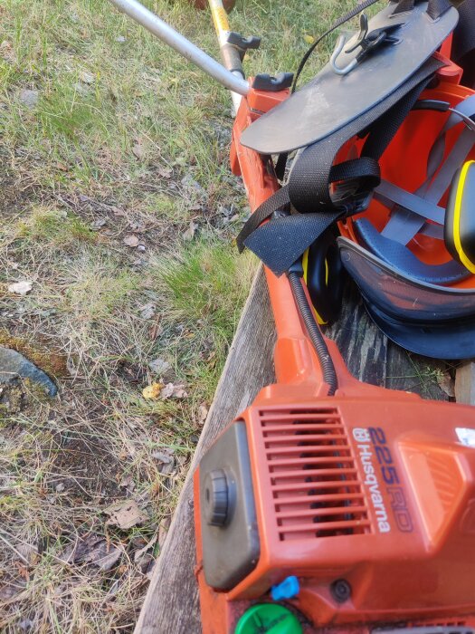 Röd motorsåg Husqvarna, svart hörselskydd, verktygsbälte, utomhus på en träbänk mot gräsbakgrund.