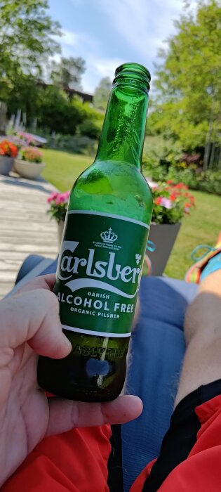 Person håller en flaska alkoholfri Carlsberg-öl ute i soligt trädgård med grönska och blommor.