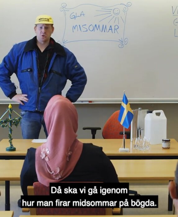 Man i keps står framför whiteboard med texten "GLAD MIDSOMMAR", lyssnande person med rosa huvudduk, svensk flagga ses.