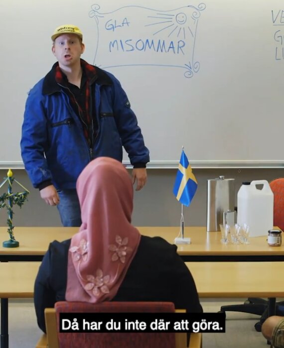 Man i blå jacka och keps framför whiteboard, svensk flagga, kvinna i hijab, text "GLAD MIDSOMMAR", svenskhet tema.