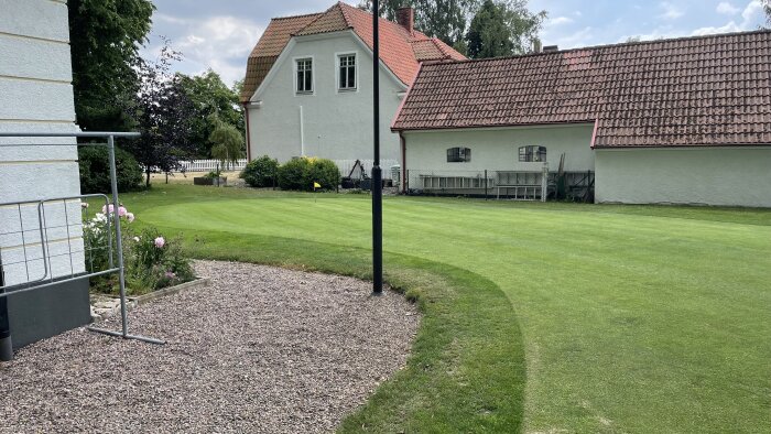 Trädgård med klippt gräs, hus, grusgång, blommor och ett staket i dagsljus.