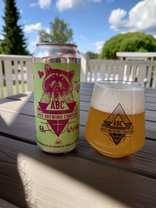 Ölburk och glas på terrassbord med trädgård i bakgrunden, soligt väder, Apex Brewing Company.