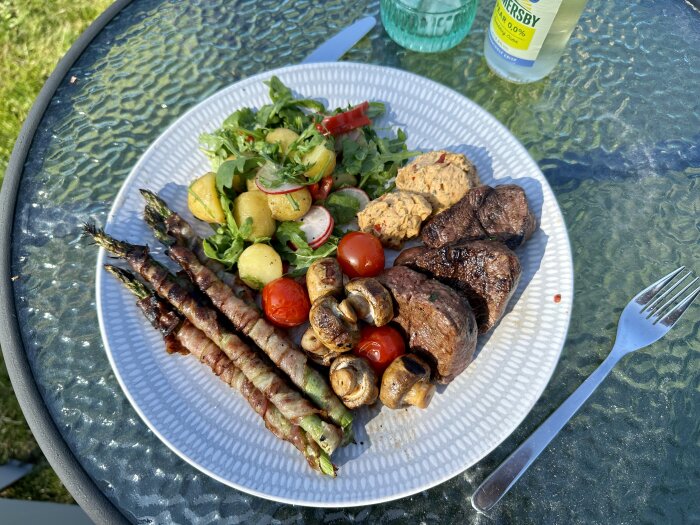 Grillat kött, sparris i bacon, sallad, champinjoner, potatis och dryck utomhus.