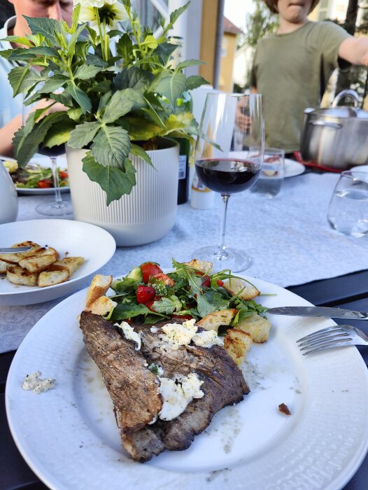 Middag uteservering med grillad köttbit, sallad, stekt potatis, rödvin, människor i bakgrunden, sommarstämning.