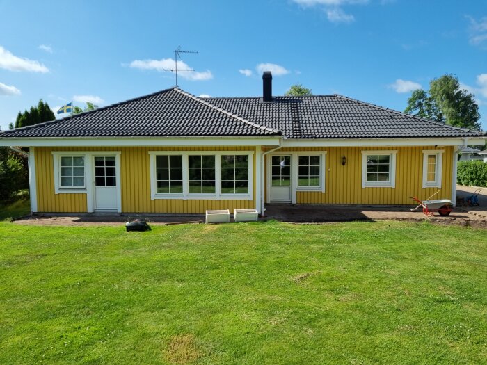 Gult hus med svart tak, vit veranda, svensk flagga, gräsmatta, kratta, skottkärra, soligt väder.