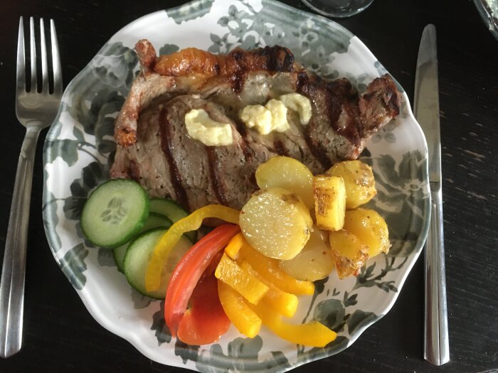 Grillad biff med smör, stekt potatis, färska gurk- och paprikaskivor på blommig tallrik.
