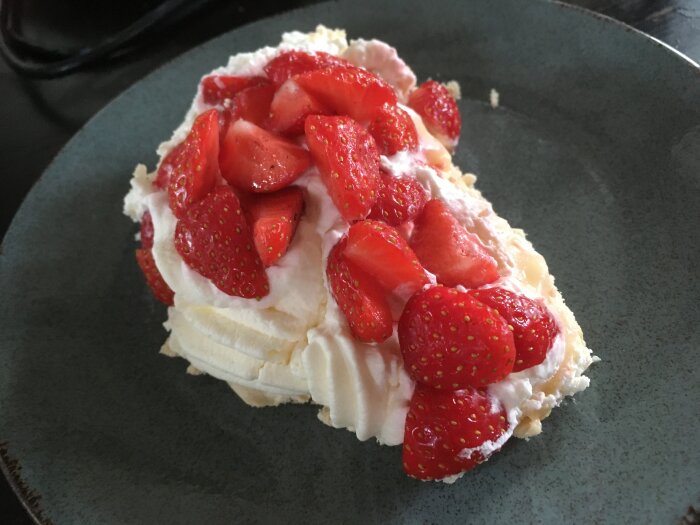 Ett stycke pavlova med grädde och skivade jordgubbar på en mörk tallrik.