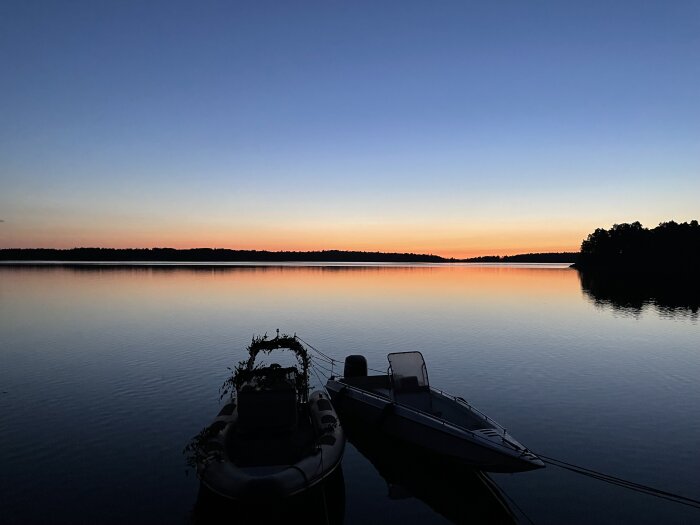 Två båtar vid sjön under solnedgången, lugnt vatten, skogssilhuett, himmel i blått och orange.