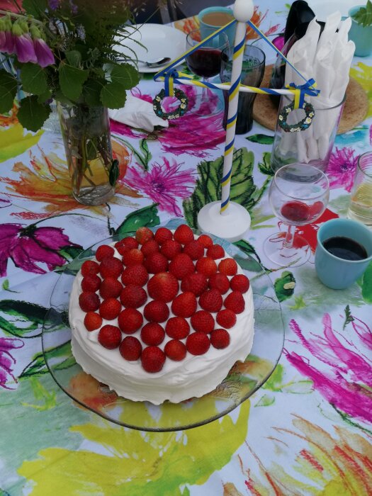 Sommarfika utomhus med jordgubbstårta, blommor, färgglad duk och midsommardekoration.