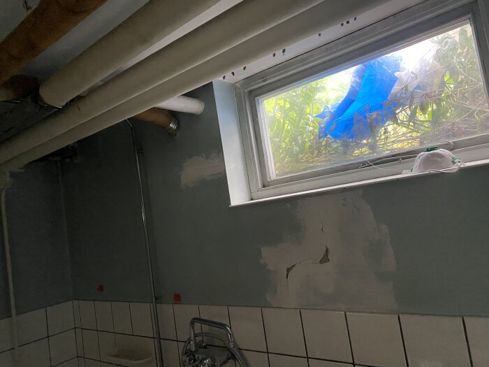 Ett badrum under renovering med otäckt fönster, synliga rör och patchad vägg.