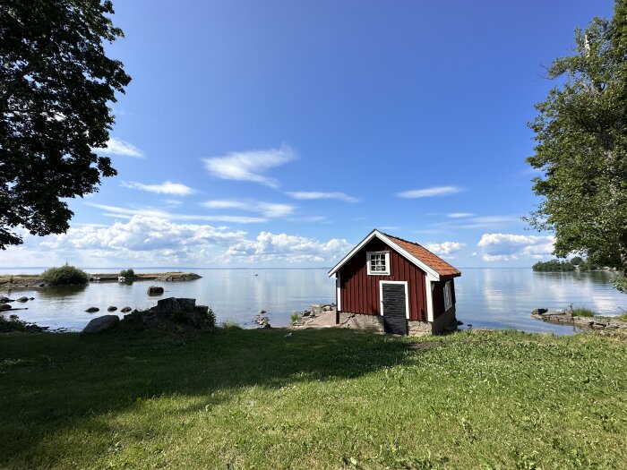 En röd stuga vid sjökanten, blå himmel med moln, grönt gräs, träd och stenar, lugnt vatten.