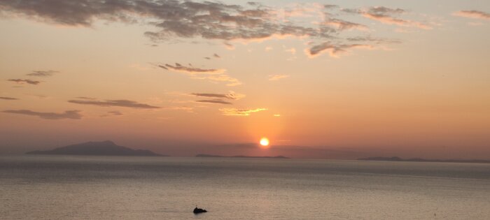 Solnedgång över havet, silhuett av berg i horisonten, moln, orange himmel, ensam båt på vattnet.