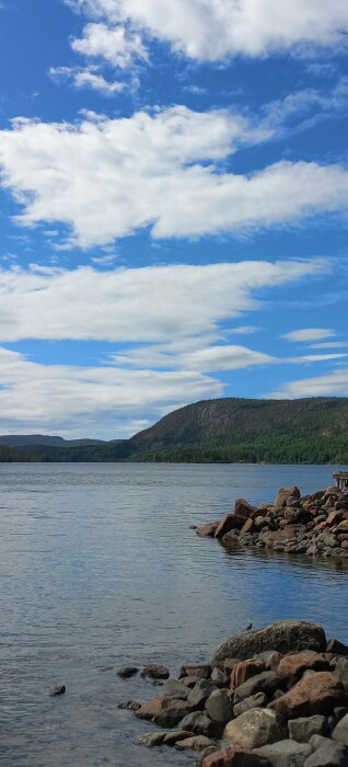 Stillhet vid en sjö med stenstrand, kullar i bakgrunden och blå himmel med vita moln.