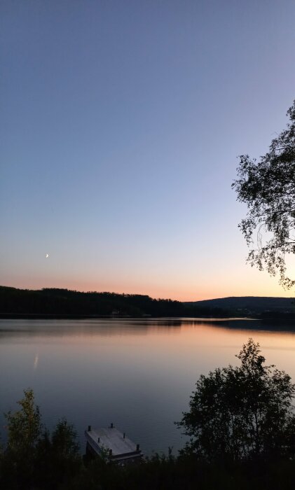 Ett stilla sjölandskap vid skymning med brygga, träd, berg i bakgrunden och en tunn månskära.