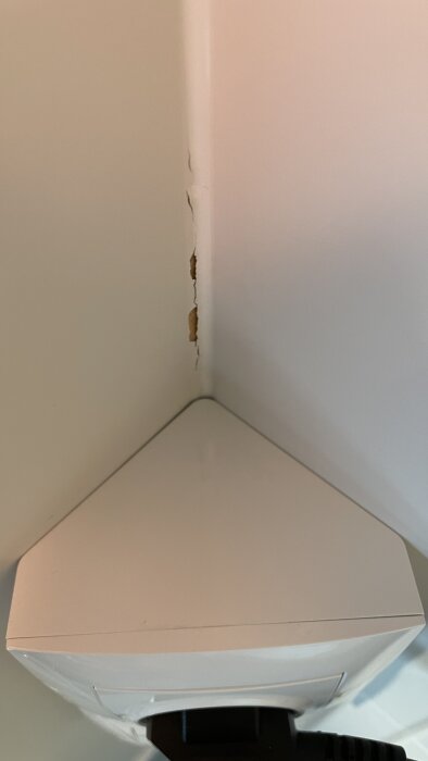 Vit hörnhylla under skadad vägghörna med sprickbildning och skalar färg.