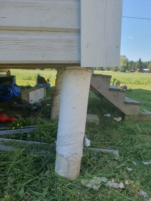 Hus på pelare, grön gräsmatta, oordning med trädgårdsredskap och byggmaterial runtomkring.